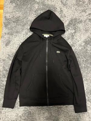 Svart hoodie från Lacoste med dragkedja - Svart hoodie från Lacoste i klassisk passform, med huva och snörning. Tröjan har dragkedja framtill, två fickor och den ikoniska gröna krokodilloggan på bröstet. Materialet är mjuk bomull, perfekt för chill dagar.