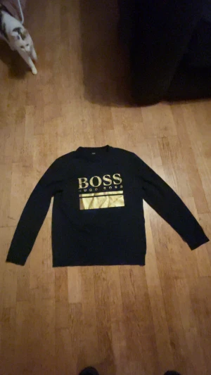 Svart långärmad tröja Hugo Boss - Svart långärmad tröja från Hugo Boss med stort guldtryck på bröstet. Snygg och stilren design med rund hals och mjukt material som känns skönt mot huden. Perfekt för dig som gillar klassiska märken och vill sticka ut med gulddetaljer.