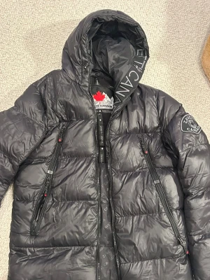 Svart dunjacka från Zavetti Canada - Svart dunjacka från Zavetti Canada med huva och coolt badge på ärmen. Jackan har quiltad design, dragkedjor med röda detaljer och snyggt mönstrat innerfoder. Perfekt för kalla dagar och har en riktigt urban vibe.