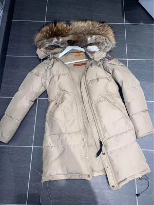 Beige Parajumpers dunjacka med päls - Säljer en beige Parajumpers dunjacka med fluffig päls på huvan. Det finns en skada på vänster arm som finns i annonsen, har använt den max tre gånger men lyckats få en skada på tyget. Utöver det är den i toppen skick, ej tappat sin form alls. 