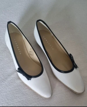 Vita ballerinaskor med blå kant Consuelo - Snygga vita ballerinaskor från Consuelo i storlek 37, med marinblå kant och liten rosett framtill. Skorna har spetsig tå och platt sula med diskret kilklack. Tillverkade i skinn med beige yttersula. Perfekt för dig som gillar klassisk och stilren design.