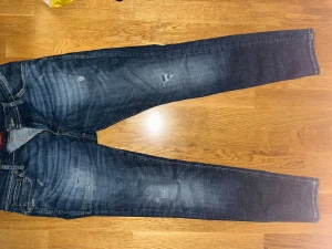 Mörkblå jeansbyxor från Jack & Jones - Snygga mörkblå jeansbyxor från Jack & Jones med klassisk femficksdesign och slitna detaljer framtill. Jeansen har en rak passform och är tillverkade i mjukt denimtyg med lätt tvättad look. Perfekta för dig som gillar en avslappnad stil.