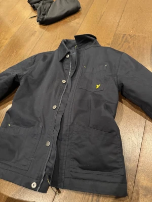 Mörkblå jacka från Lyle & Scott - Snygg mörkblå jacka från Lyle & Scott med klassisk krage, stora fickor framtill och gul logga på bröstet. Jackan har både dragkedja och knappar, samt en stilren design som passar perfekt till vardagslooken.