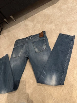 Replay Anbass jeans blå storlek 33  - Säljer ett par snygga Replay Anbass jeans i blå tvätt med slitningar och klassiska fem fickor. Jeansen har normal passform och raka ben, med detaljerad söm på bakfickorna och läderpatch med Replay-logga i midjan. Perfekt för dig som gillar stilrena jeans.