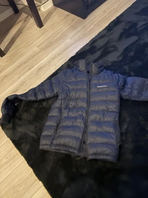 Mörkblå dunjacka från Peak Performance - Säljer en mörkblå dunjacka från Peak Performance med horisontella quiltade paneler och dragkedja framtill. Jackan har två sidofickor med dragkedja och en hög krage. Perfekt för kalla dagar och har en clean, sportig look. Och har ett litet hål i en av armarna 