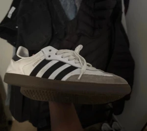 Adidas Samba sneakers vit/svart - Adidas Samba sneakers i klassisk vit och svart färg med bruna gummisulor. Skorna har de ikoniska tre svarta ränderna på sidan, snörning framtill och rund tå. Ovandelen är i skinn och mocka, med svart detalj på hälen. Perfekt för dig som gillar retrostil och streetwear.