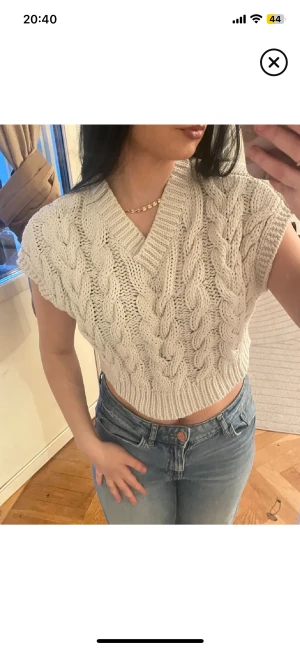 Beige stickad croppad topp - Säljer en beige stickad croppad topp med flätstickat mönster och v-ringad hals. Toppen är ärmlös och har en loose passform som ger en chill vibe. Perfekt att styla med jeans eller kjol för en trendig look. Strl 134/140 men passar fint på mig som är xs/s