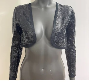 Svart paljett bolerotopp från C - Glittrig svart bolerotopp från C, täckt med paljetter för maxad partykänsla. Croppad modell med lång ärm och öppen front, perfekt att slänga över en klänning eller topp för extra bling. Snygg och trendig detalj till din outfit.