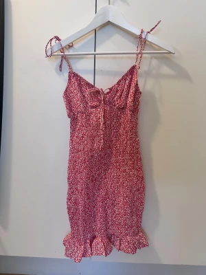 Röd blommig kortklänning med volang - Supersöt röd kortklänning med små vita blommor, knytband på axlarna och volangkant längst ner. Klänningen har en feminin v-ringning med knyt framtill och är ärmlös, perfekt för varma dagar. Materialet känns lätt och luftigt.