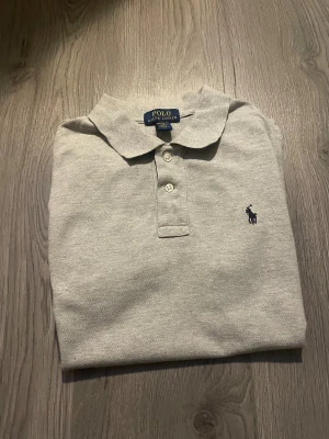 Grå piké från Polo Ralph Lauren - Snygg grå pikétröja från Polo Ralph Lauren med klassisk krage och tre vita knappar framtill. Tröjan har korta ärmar och den ikoniska broderade loggan i mörkblått på bröstet. Tillverkad i mjuk bomull som är skön mot huden. Hör av dig fundeing. Snabb köp bättre pris :)