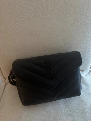 Svart  axelväska YSL - Säljer en svart quiltad axelväska från YSL med klassisk YSL-logga framtill. Perfekt för dig som vill ha en lyxig och tidlös accessoar. Skriv privat för fler frågor. Den har nån pytte pytte liten skråma man knappt ser. Kan skicka bild. Pris går diskutera vid snabb affär 