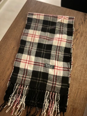Rutig halsduk från Burberrys i ull - Klassisk rutig halsduk från Burberrys i 100% ull. Halsduken har fransar i ändarna och den ikoniska Burberrys-etiketten. Perfekt accessoar för att lyfta din outfit. Anledningen att de stör ”burberrys” är för att de är en äldre modell.