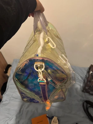 Louis Vuitton holografisk handväska - Säljer en unik holografisk handväska från Louis Vuitton med genomskinligt material och ikoniskt LV-mönster. Väskan har vita detaljer, robusta handtag och en cool kedja i plast. Perfekt för dig som vill sticka ut med en futuristisk look.