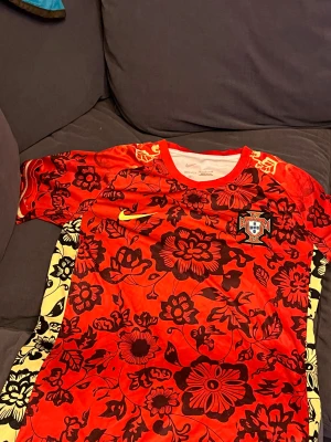 Fotbollströja - Säljer en unik Portugal fotbollströja från Nike i röd färg med blommigt mönster i svart och detaljer i gult längs sidorna. Kortärmad modell med rund hals och broderat Portugal-emblem på bröstet. Materialet är lätt och andas, perfekt för match eller träning.