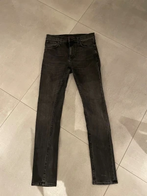 Nudie jeans grå/svart - Tja säljer nu ett par feta grå/Svarta jeans från Nudie  med slim passform och klassisk femficksdesign. Jeansen har svarta sömmar, knapp och dragkedja framtill  Perfekt för dig som gillar en clean och stilren look. Skicket är helt perfekt som nytt ungefär och storleken är w28 l32 passar dig som är runt 170-183cm lång skulle jag tro!! Har du några övriga frågor är de bara att höra av sig🙌🏼mvh em_resell🤩