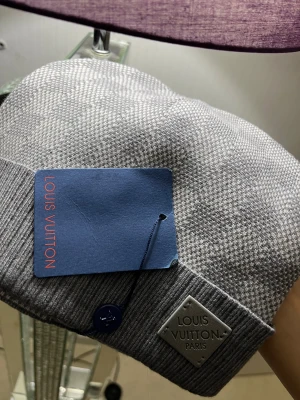 Grå stickad mössa  - Snygg grå stickad mössa  med diskret rutmönster och ribbad kant. Märkets metallbricka framtill och blå etikett på insidan. Mjuk och lyxig känsla, perfekt för dig 