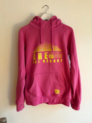 Rosa hoodie från United by Altitude - Säljer en snygg rosa hoodie från United by Altitude med gul tryckt text 'Åre Ski Resort' och logga på magen. Tröjan har huva med snörning. Perfekt för dig som gillar färg och vill sticka ut. Finns en fläck på fickan, se bild.