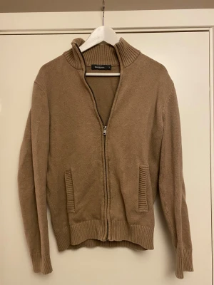 Beige stickad kofta från Dressmann - Säljer en beige stickad kofta från Dressmann i storlek M. Koftan har ribbad krage, muddar och fickor samt dragkedja framtill. Perfekt för dig som gillar enkel och stilren design med lite extra struktur i detaljerna.