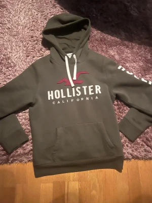  Hollister hoodie  - Exklusiv olivgrön hoodie från Hollister med stor vit logga och röd fågel på bröstet samt Hollister-tryck längs ena ärmen. Materialet är bomullsblandning. 10/10 skick, knappt använd. Perfekt för dig som vill ha en stilren och snygg hoodie till vintern. Skriv vid intresse