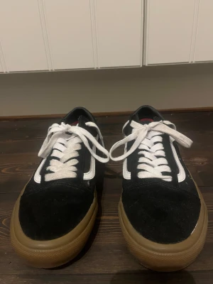 Vans Old Skool svart/vit sneakers - Säljer ett par Vans Old Skool sneakers i svart och vitt med klassisk sidestripe och gummisula i brun. Skorna har snörning med vita skosnören, rund tå och platt sula. Ovandelen är i mocka och canvas, perfekt för dig som gillar skate och streetstyle.