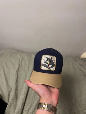 Tom & Jerry keps från Capslab - Snygg Tom & Jerry keps från Capslab med beige skärm och mörkblå front. Framsidan har en patch med Tom och mesh på sidorna för extra ventilation. Broderade loggor på sidan och insidan, justerbar passform. Perfekt för dig som gillar retro och tecknade serier.