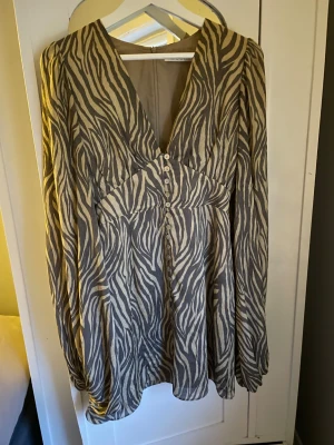 Zebramönstrad kortklänning från Adoore - Snygg kortklänning från Adoore i beige och grått zebramönster. Klänningen har v-ringning, långa ärmar med knappar vid manschetten och dekorativa knappar framtill. Materialet är lätt och ger ett flowy intryck. En knapp saknas längst ner, vilket är synd då den är använd två gånger🥲