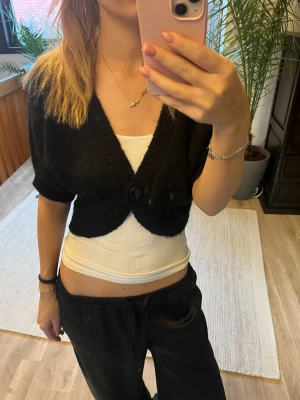 Svart croppad kofta från H&M - Svart croppad kofta från H&M med korta ärmar och djup v-ringning. Koftan har en stor knapp framtill och är stickad i mjukt material. Perfekt att slänga över ett linne för en chill look.