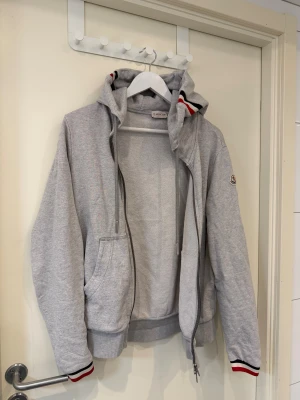 Grå hoodie från Moncler - Snygg grå hoodiejacka från Moncler med dragkedja, huva och snörning. Hoodien har ribbade muddar med röd, vit och svart rand samt Moncler-logga på ärmen. Perfekt för chill dagar och streetwear-stil. Köpt från garmsmarket. Kom privat för fler frågor och bilder.