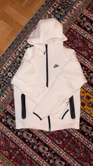 Vit hoodie från Nike med svarta detaljer - Säljer en clean vit hoodie från Nike med svarta dragkedjor och logga på bröstet. Tröjan har huva, långa ärmar och praktiska fickor med dragkedja. Perfekt för dig som gillar sportig och stilren look. Materialet är mjukt och känns skönt mot huden.