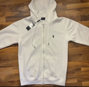 Vit hoodie från Polo Ralph Lauren - Vit hoodie från Polo Ralph Lauren med dragkedja och klassisk logga broderad på bröstet. Tröjan har huva med snörning, två fickor fram och ribbade muddar. Perfekt för chill dagar och enkel att matcha med jeans eller joggers.