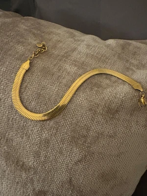 Guldigt platt armband med kedja - Snyggt guldigt armband med platt, fiskbensliknande design och justerbar kedja. Armbandet har en elegant och modern look som passar perfekt till din stil. Lätt att bära och matcha med andra smycken. Perfekt accessoar för dig som gillar guld och stilrena detaljer. Rostfritt