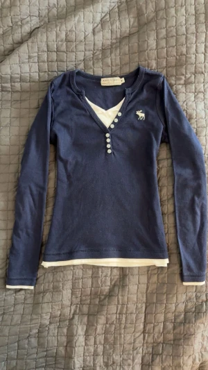 Abercrombie & fitch mörkblå långärmad tröja  - Mörkblå långärmad tröja från Abercrombie & Fitch med vit logga på bröstet och dekorativa knappar vid v-ringningen. Storlek M men passar en Xs. Tröjan har en vit kant vid halsringning och nederkant som ger en layered look. 