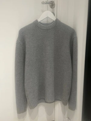 Grå stickad tröja från Zara - Säljer en stilren grå stickad tröja från Zara i storlek M. Tröjan har en rundad hals och ribbade muddar vid ärmslut och nederkant. Perfekt för dig som gillar enkel och clean look med mjukt material.