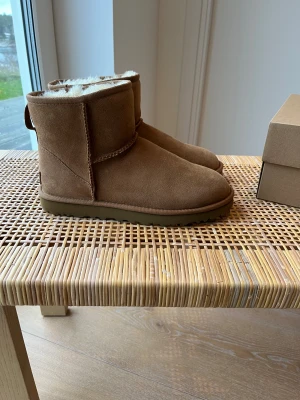 UGG Classic Mini II –  Chestnut. - Mysiga bruna UGG boots i mocka med mjukt vitt foder och platt sula. Klassisk rund tå och kort skaft, perfekta för kalla dagar. UGG-logga bak på hälen och grov yttersula för bra grepp. Dom är endast använda 2 gånger, alltså är dom i helt nytt skick. Inget tecken på att dom har använts. Nypris är 2 200 kr.  Storlek 39.