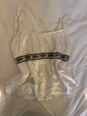 Vit sidenlinnetopp med svart spets - Supersöt vit linnetopp i glansigt sidenmaterial med tunna axelband. Toppen har en svart spetsdetalj med broderade rosor. Perfekt för dig som gillar romantiska och feminina detaljer. Sval och lätt, passar till många olika stilar.