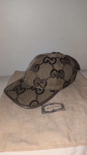 Beige Gucci jumbo keps  - Snygg beige keps från Gucci med det klassiska GG-logomönstret i mörkbrunt. Kepsen har justerbar rem baktill och är tillverkad i bomullsmaterial med detaljer i skinn. Perfekt för dig som vill ha en lyxig och trendig accessoar. Strl M 58cm byten funkar och skriv vid frågor 