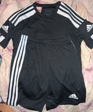 Svarta Adidas Shorts och T-shirt  - Säljer ett par svarta Adidas Shorts och T-shirt med klassiska vita ränder längs sidorna och Adidas-logga framtill. De har elastisk midja och är gjorda i ett lätt och skönt polyestermaterial, perfekta för träning eller sport. Snygg och sportig look som alltid funkar.