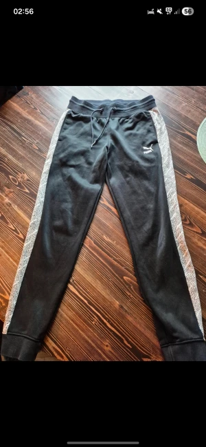 Svarta mjukisbyxor från Puma M - Säljer ett par svarta mjukisbyxor från Puma i storlek M. Byxorna har vita och svarta mönstrade sidopaneler, resår i midjan med snörning och ribbade muddar vid bensluten. Puma-logga på vänster lår. Perfekta för chill eller träning.