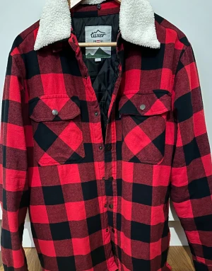 Rutig jacka från Tuxer med teddykrage - Röd och svart rutig jacka från Tuxer med vit teddykrage. Jackan har två bröstfickor med knappar, quiltat svart innerfoder och tryckknappar framtill. Perfekt för lager-på-lager  och chill höst/vinter.