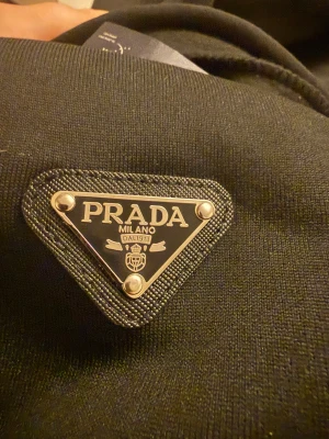 Svart hoodie från Prada med dragkedja - Svart hoodie från Prada med ikonisk metalltriangel på bröstet, justerbar huva med snörning och praktiska fickor framtill. Tröjan har hel dragkedja och är tillverkad i mjukt material som känns lyxigt. Perfekt för dig som gillar exklusiva streetwear-vibbar. Passar även storlek M.