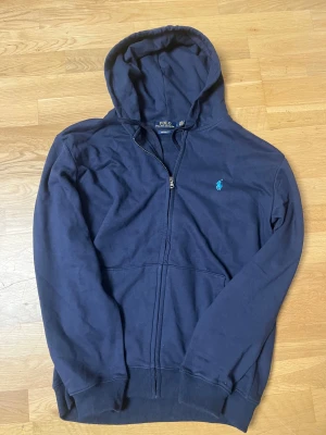 Marinblå hoodie från Polo Ralph Lauren - Snygg marinblå hoodie från Polo Ralph Lauren i storlek L. Tröjan har dragkedja framtill, huva med snörning och det klassiska Polo-logot broderat på bröstet. Tillverkad i mjuk bomull och har långa ärmar samt ribbade muddar vid ärmslut och nederkant.