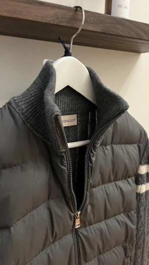  Moncler Cardigane  - Snygg grå pufferjacka från Moncler med stickade, kabelmönstrade ärmar i ull och vita ränder. Jackan har hög ribbad krage, tvåvägsdragkedja och Moncler-logga på ärmen. Perfekt för dig som vill ha en unik och varm jacka med coola detaljer.
