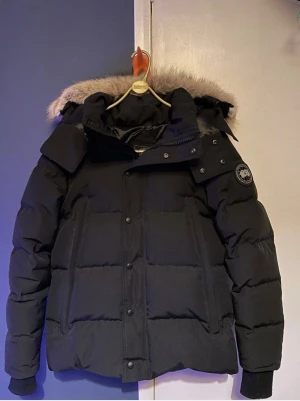 Svart pufferjacka Canada Goose - Svart pufferjacka från Canada Goose med fet päls på huvan och klassisk logga på ärmen. Jackan har dragkedja och knappar framtill. Perfekt för kalla vinterdagar och riktigt snygg till streetwear.