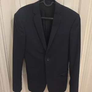 Mörkblå kavaj från Filippa K - Snygg mörkblå kavaj från Filippa K i klassisk modell med smal krage och enkel knappstängning. Kavajen har två fickor framtill och en bröstficka. Den är använd 1 gång så den är i ett otroligt bra skick!