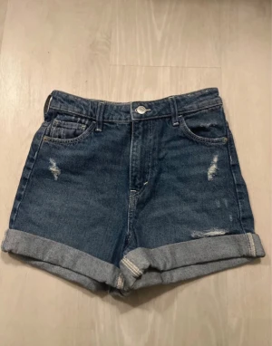 Blå denimshorts high waist &Denim - Snygga blå denimshorts från &Denim med avslappnad passform och hög midja. Shortsen har slitna detaljer, klassiska fem fickor och uppvikta benslut. Perfekta för dig som gillar en chill och trendig stil under varma dagar.