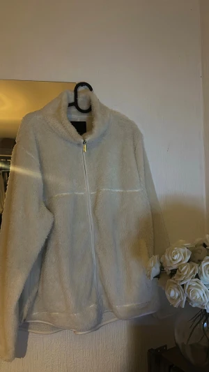 Beige teddyjacka med dragkedja - Mysig beige teddyjacka med hög krage och hel dragkedja framtill. Jackan har en fluffig och mjuk känsla, perfekt för kyliga dagar. Enkel och stilren design utan synliga loggor eller mönster. Passformen är normal och ärmarna är långa.