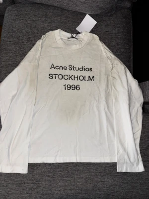 Vit långärmad tröja Acne Studios S - Vit långärmad tröja från Acne Studios i storlek S, med tryck 'Acne Studios STOCKHOLM 1996' på bröstet. Tröjan är tillverkad i 100% bomull och har en klassisk rund hals. Perfekt för dig som gillar minimalistisk och stilren design.