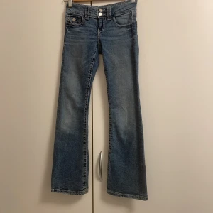 Blå bootcut jeans från Gina Tricot - Lågmidjade snygga blå jeans från Gina Tricot i storlek 146 med bootcut-ben och klassisk femficksdesign. Jeansen har dubbla silverknappar i midjan, markerade sömmar och en cool tvättad look. Perfekta för dig som gillar retrovibbar och bekväm stil.