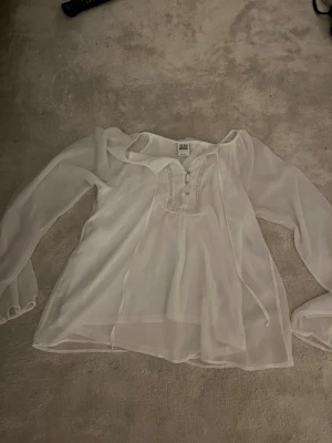 Vit blus med spets från Vero Moda - Säljer en superfin vit blus från Vero Moda i transparent, lätt polyester. Blusen har långa ärmar med resår vid ärmslut, spetsdetaljer runt knäppningen och tre små knappar framtill. Omlottkänsla och knytband vid halsen ger en romantisk vibe.