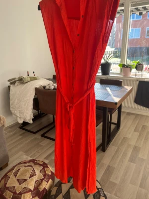 Röd långklänning från Billabong - Snygg röd långklänning från Billabong med knappar hela vägen framtill och knytskärp i midjan. Klänningen är ärmlös och har en lätt, luftig känsla – perfekt för varma dagar. Materialet är skrynklat och ger en avslappnad vibe.